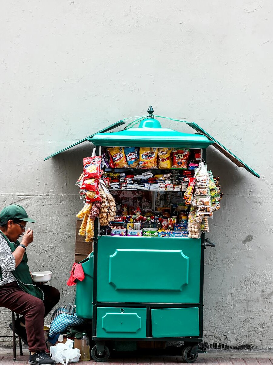 A woman enjoying a meal while sitting next to her snack cart on a city street.